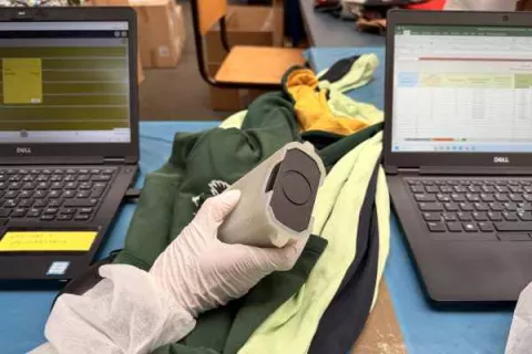 Hand mit Messgerät analysiert Textilien während einer Untersuchung zur Ökobilanzierung, begleitet von Datenauswertung auf Laptops.